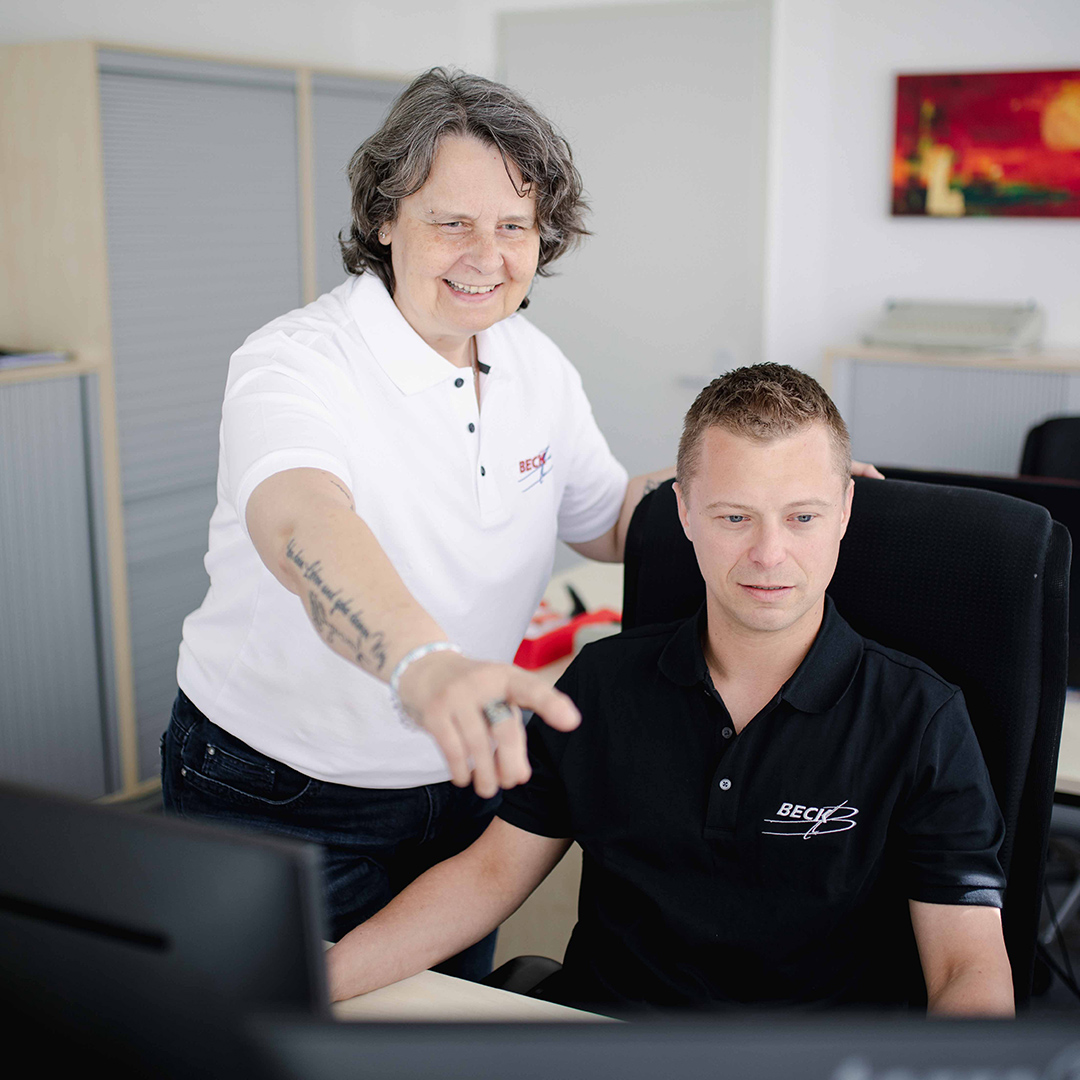 Stehende Frau an einem Schreibtisch zeigt dem daneben sitzenden Mann am Schreibtisch etwas auf dem Computer.