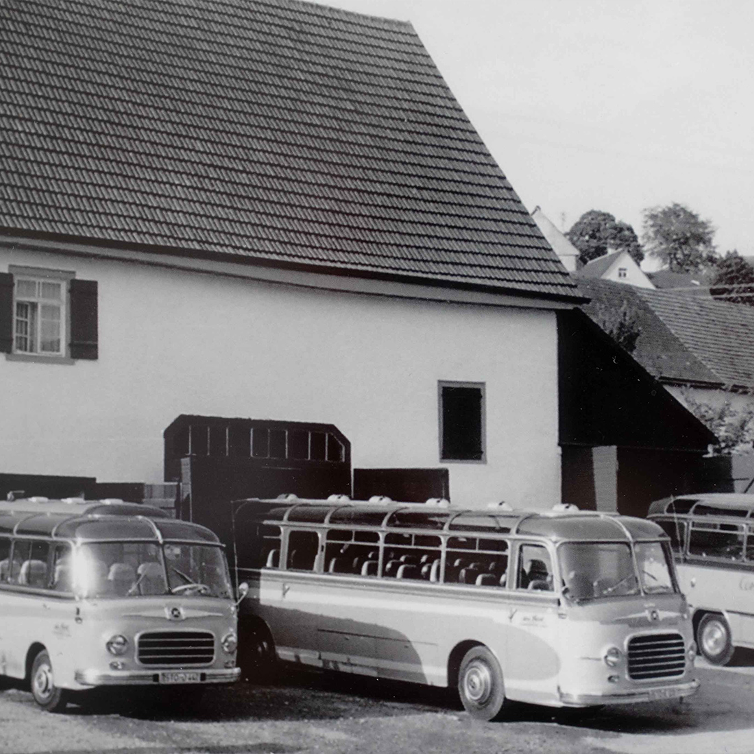 Ein Schwarz-Weiß Bild von sehr alten Bussen die vor einem größeren Gebäude mit Garage parken.