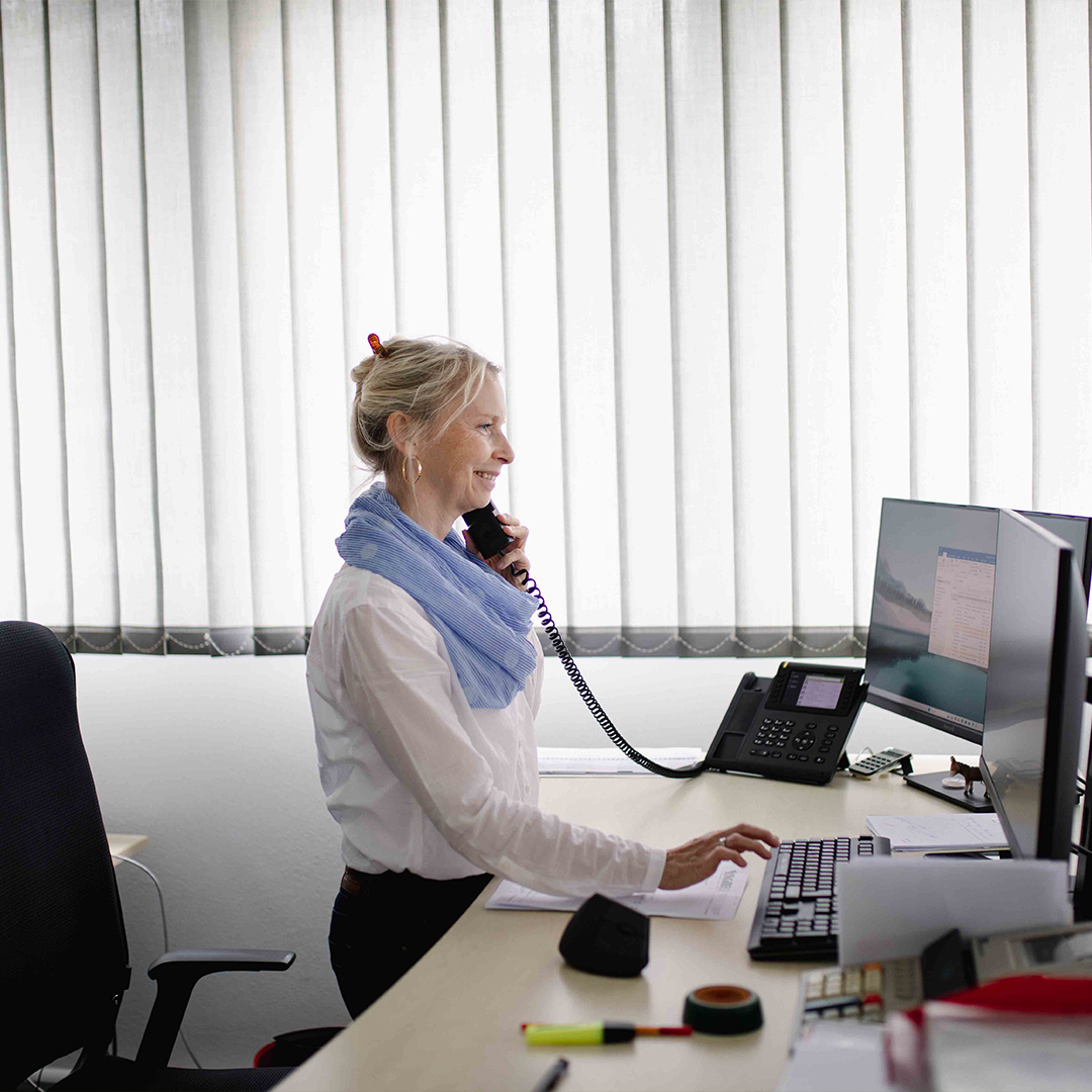 Frau steht an ihrem Schreibtisch, schaut in den Computer und telefoniert dabei.