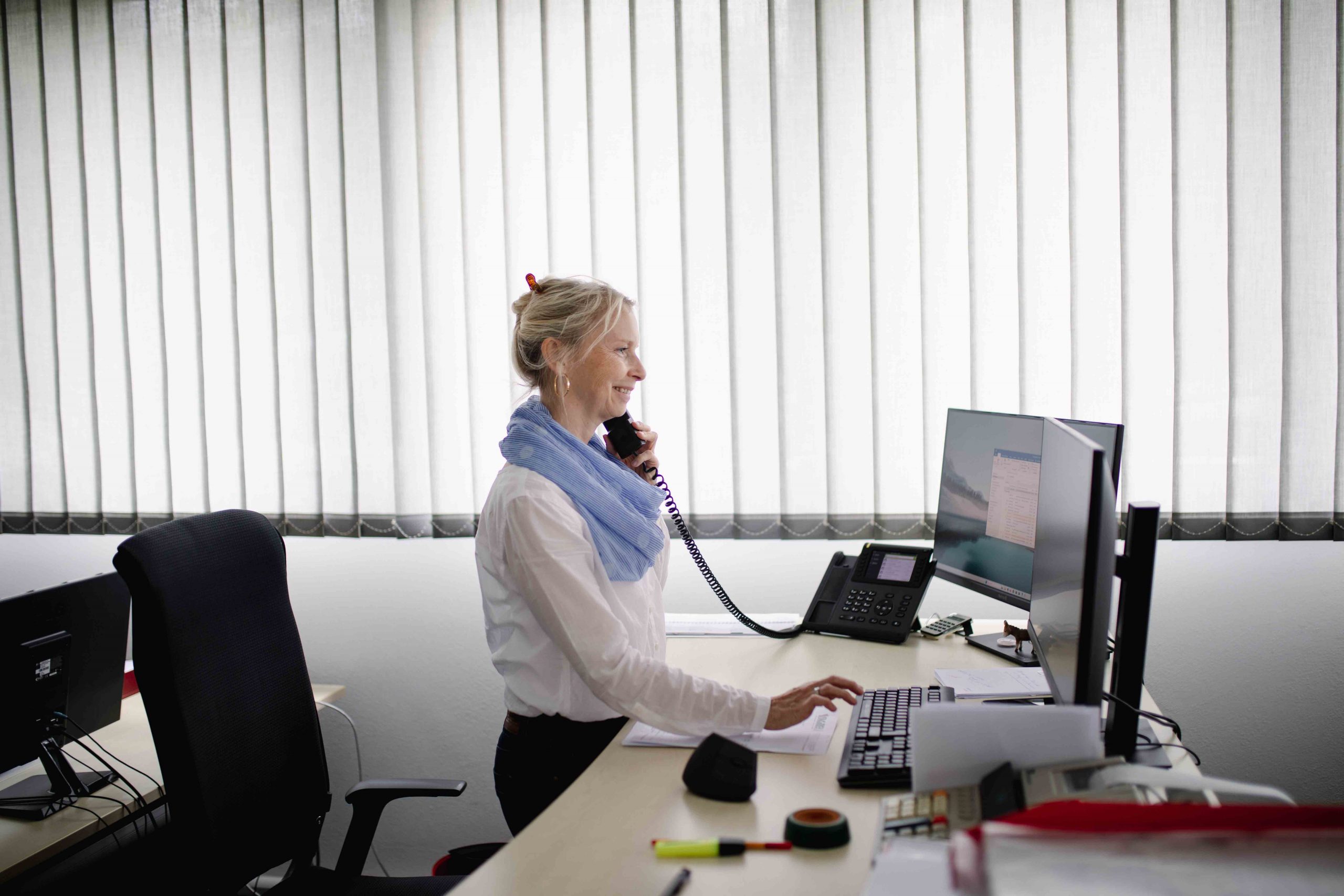 Frau steht am Schreibtisch, schaut in den Computer und telefoniert dabei.